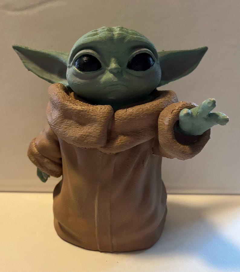 May include: A figurine of a character with large black eyes, green skin, and large ears. The character is wearing a brown robe and has one hand raised. The figurine is a collectible item.