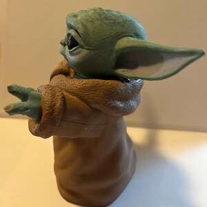 May include: A figurine of a character with large ears and green skin, wearing a brown robe. The figure has its arms outstretched, with three fingers visible on each hand. The character's eyes are black, and the figure is set against a neutral background.