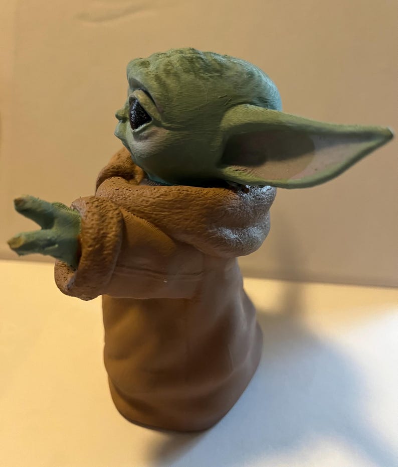 May include: A figurine of a character with large ears and green skin, wearing a brown robe. The figure has its arms outstretched, with three fingers visible on each hand. The character's eyes are black, and the figure is set against a neutral background.