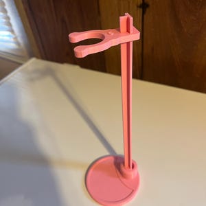 May include: A pink plastic doll stand with a round base and a hook for holding a doll.