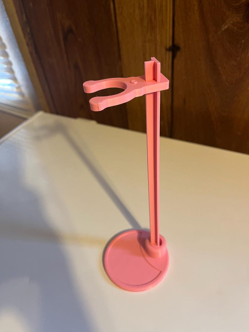 May include: A pink plastic doll stand with a round base and a hook for holding a doll.