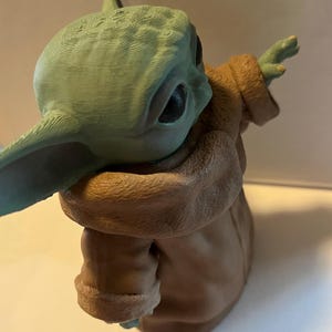 May include: A green and brown figurine of a character with large ears and a tan robe. The figurine has a textured surface and is posed with one arm outstretched. The character's large eyes are visible.