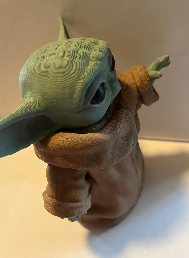 May include: A green and brown figurine of a character with large ears and a tan robe. The figurine has a textured surface and is posed with one arm outstretched. The character's large eyes are visible.