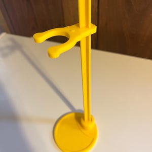 May include: A yellow plastic doll stand with a round base and a hook for holding a doll.