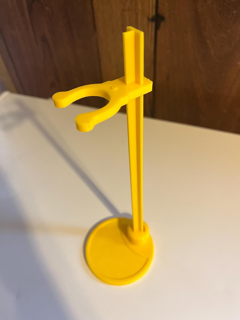 May include: A yellow plastic doll stand with a round base and a hook for holding a doll.