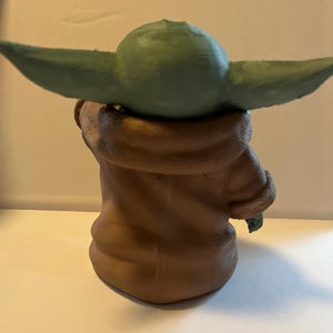 May include: A green-skinned character figurine with large ears, wearing a brown robe with a furry collar. The figurine is a stylized representation of a popular science fiction character.