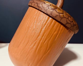 May include: A large, acorn-shaped container with a textured, brown body and a detailed, brown cap. The cap features a lighter brown, textured pattern and a small, brown stem. The container is set against a white surface and a dark blue background.