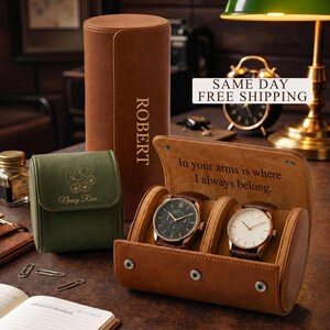 May include: A tan leather watch roll case with two wristwatches inside, featuring the engraved text 'In your arms is where I always belong.' Next to it is a tall tan leather case labeled 'ROBERT' and a small green leather case with a zodiac graphic.