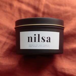 Puede incluir: Una lata de vela negra con borde dorado y etiqueta blanca. La etiqueta dice "nilsa" en una fuente moderna, con "ORGANIC SOY WAX CANDLES HAND POURED IN PHILADELPHIA" debajo. La vela está sobre una tela color óxido.
