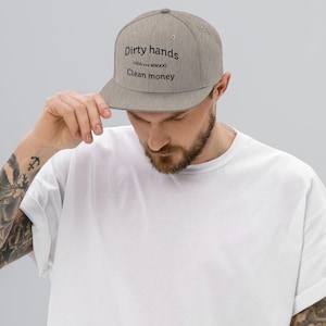 Può includere: Un cappellino da baseball grigio a tesa piatta con la scritta "Dirty hands" sopra "Clean money" in nero. Il cappellino è indossato da una persona che indossa una maglietta bianca.