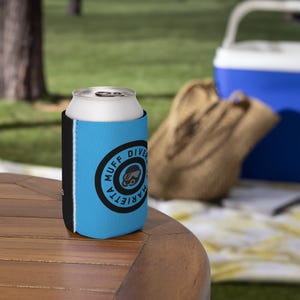 Può includere: Un portabevande blu e nero con la scritta "MUFF DIVER MARIETTA" in un design circolare. Il portabevande contiene una lattina argentata. Lo sfondo mostra una scena da picnic con una borsa frigo e una borsa.