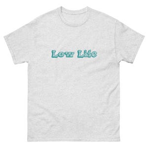 Può includere: T-shirt grigio chiaro con la scritta "Low Life" in turchese, bordata di un turchese più scuro. La maglietta ha un girocollo e maniche corte. Il testo è centrato sul petto.
