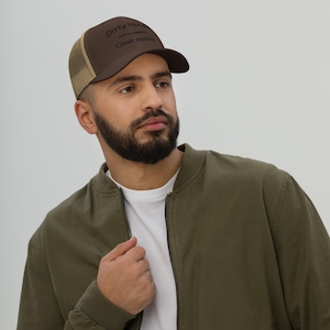Può includere: Un cappellino da camionista marrone e beige con la scritta "Dirty hands, Clean money" stampata sul davanti. Il cappello ha una visiera curva e un retro in rete. La persona indossa una giacca bomber verde.