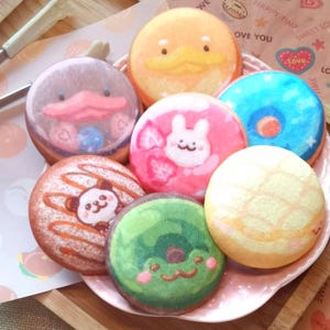 Pode incluir: Uma seleção de donuts decorados com desenhos de desenhos animados, incluindo um pato, um coelho, um panda e um sapo. Os donuts são dispostos em um prato rosa. O fundo é de cor clara.