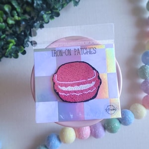 May include: An iron-on patch featuring a pink embroidered macaron with white filling. The patch is packaged in a clear plastic sleeve and is labeled "IRON-ON PATCHES". The background is a pastel color scheme.
