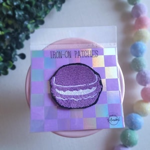 May include: A purple embroidered iron-on patch in a clear plastic package. The patch is shaped like a macaron with white filling. The package has the words "IRON-ON PATCHES" printed on it. The background is a checkered pattern.