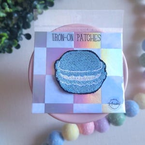 Peut inclure: Un écusson thermocollant brodé bleu en forme de macaron, avec des détails de couture blancs. L'écusson est emballé dans une pochette plastique transparente avec l'inscription "IRON-ON PATCHES". Le fond est un motif à carreaux pastel.