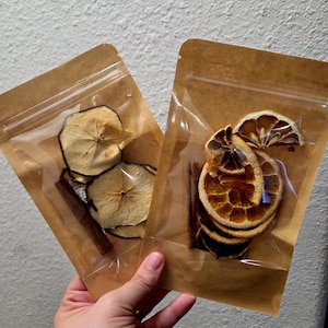 Może przedstawiać: Dwa przezroczyste plastikowe woreczki wypełnione suszonymi owocami i przyprawami. Jeden woreczek zawiera plasterki jabłka i laskę cynamonu. Drugi woreczek ma suszone plasterki pomarańczy i laski cynamonu. Woreczki trzymane są w dłoni.