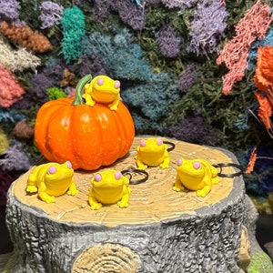 May include: Four yellow frog keychains with pink eyes are arranged on a gray tree stump with a small orange pumpkin on top. The tree stump is surrounded by green grass.