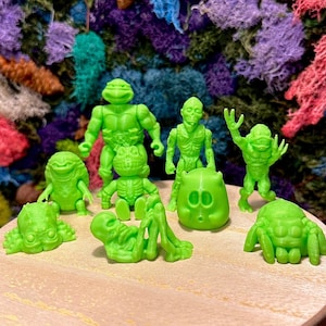 May include: A collection of small, bright green monster figurines displayed on a small wooden table. The figures include various monster designs, such as a turtle-like creature, a skeleton, and a spider. The background features a colorful array of moss.