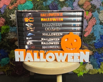 May include: A stack of DVD cases featuring the Halloween franchise, including Halloween II, Halloween Resurrection, and Halloween H20. A 3D orange pumpkin with a black face is on top of the stack. A white 3D word "HALLOWEEN" is in front of the DVD cases.