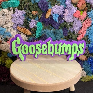 May include: A green and purple 3D printed sign that reads "Goosebumps" on a small wooden stool with black legs. The sign is sitting on a bed of colorful moss.