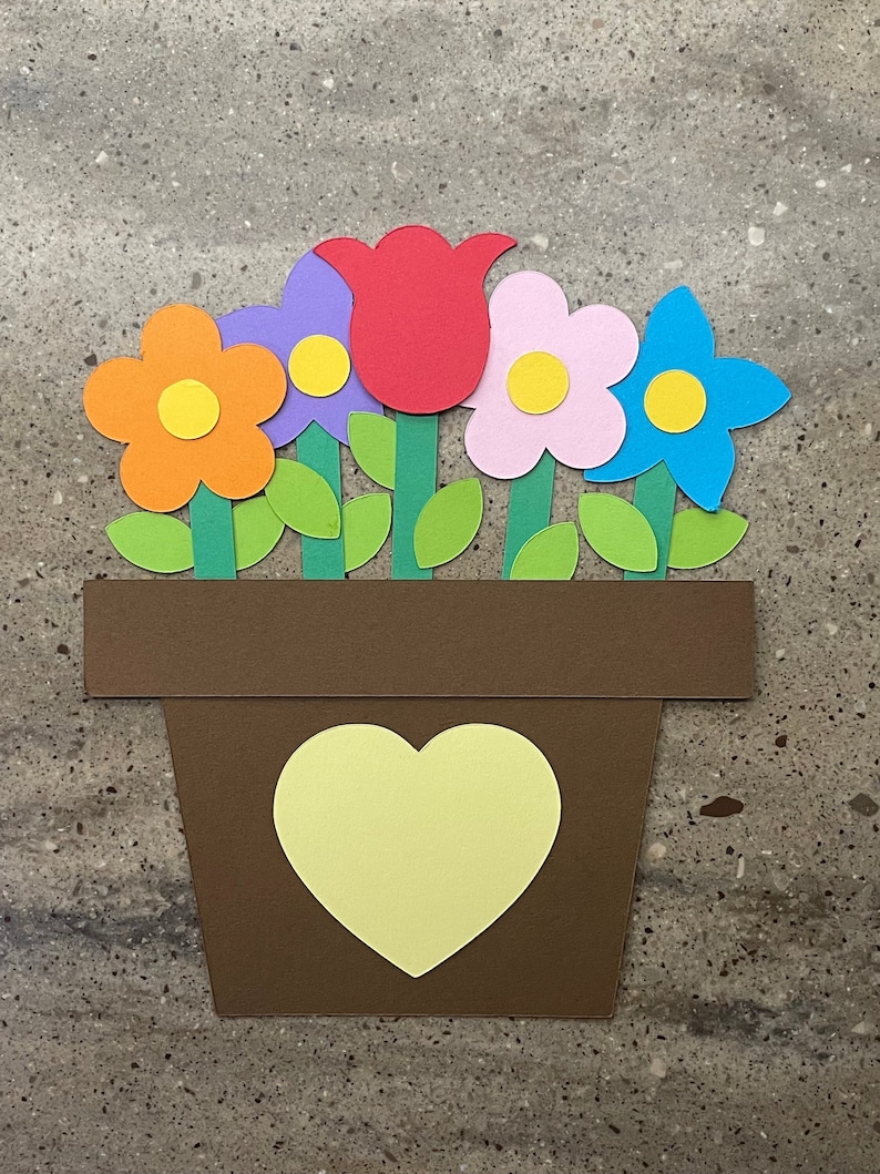 May include: A brown paper flower pot with a yellow heart on the front. The pot is filled with five paper flowers: a red tulip, a purple flower, an orange flower, a pink flower, and a blue flower. Each flower has a yellow center.