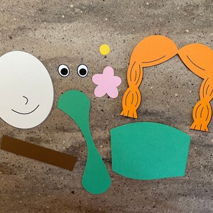 May include: A set of paper craft cutouts for a craft project. The cutouts include a white face with a smile, two black eyes, a pink flower, a brown stick, a green leaf, a green chair, and a pair of orange pigtails.