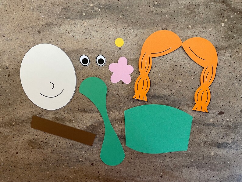 May include: A set of paper craft cutouts for a craft project. The cutouts include a white face with a smile, two black eyes, a pink flower, a brown stick, a green leaf, a green chair, and a pair of orange pigtails.