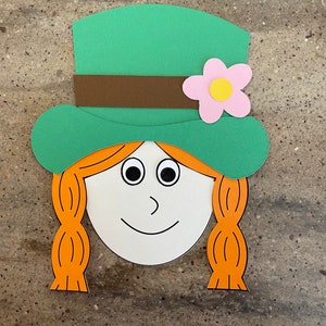 May include: A paper craft of a leprechaun face with a green hat, brown band, pink flower with a yellow center, and orange hair.