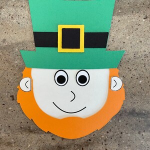 May include: A paper craft leprechaun face with a green hat, black belt, yellow buckle, white face, black eyes, orange beard, and white ears.