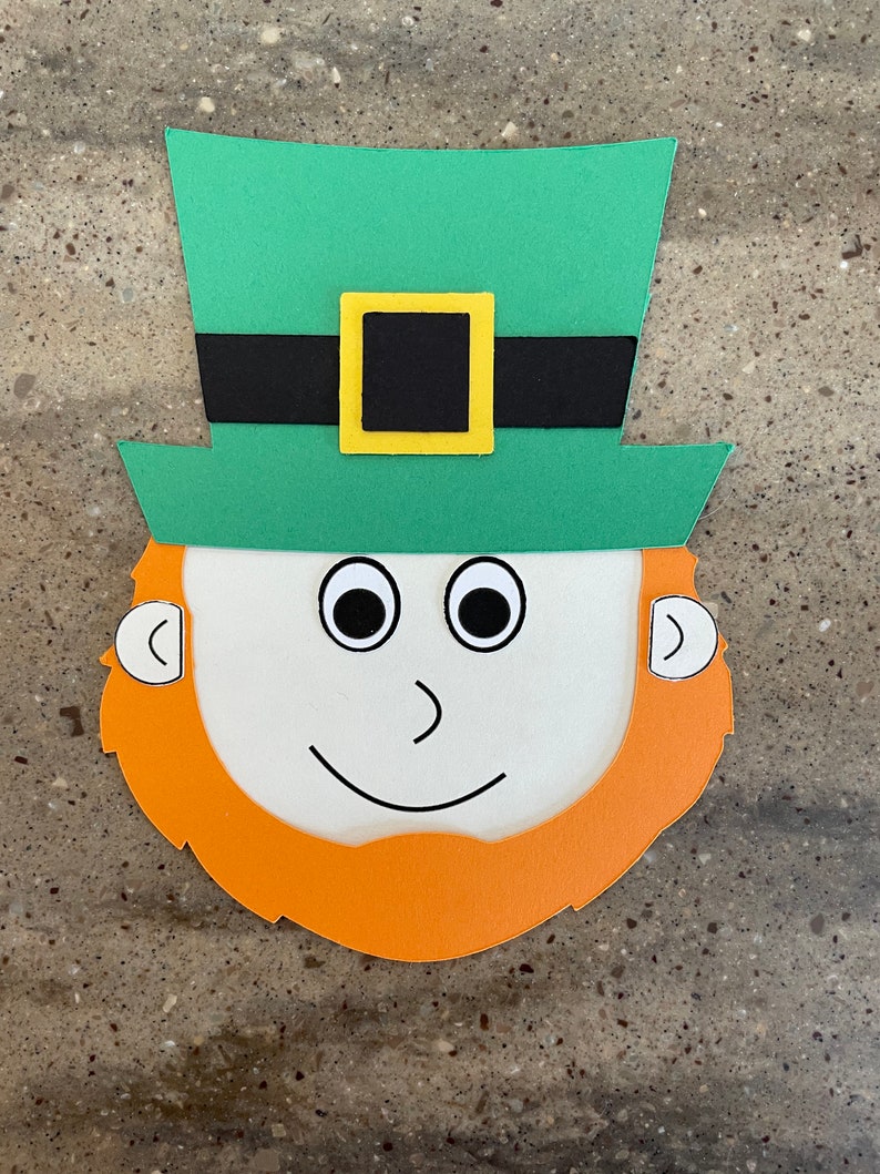 May include: A paper craft leprechaun face with a green hat, black belt, yellow buckle, white face, black eyes, orange beard, and white ears.