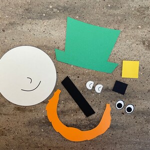 May include: A craft project with paper cutouts for making a leprechaun. The pieces include a white circle for the face, a green hat, a black mustache, orange beard, yellow square, black square, and two white eyes.