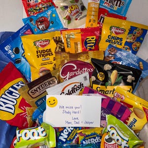 May include: A variety of snack food packages including M&M's, Keebler cookies, Ritz crackers, Snyder's pretzels, Goldfish, Smartfood popcorn, Bugles, Nerds, Pringles, and Rice Krispies Treats. A white note reads: We miss you! Study hard! Love, Mom, Dad & Jasper.