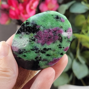 May include: A polished, oval-shaped gemstone with a vibrant mix of colors. The stone features a base of deep green, speckled with black and patches of rich, dark red. The gemstone is held in a hand, with a blurred floral background.