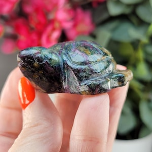 May include: A small, carved turtle figurine with a polished, iridescent surface. The turtle's shell displays a mix of dark green, purple, and gold hues. The figurine is held in a hand, with a bright orange painted fingernail visible.