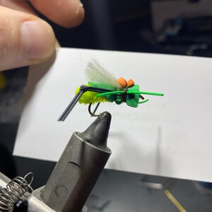 May include: A green and yellow fly fishing lure with white fur and orange accents. The lure is tied on a hook and is attached to a vise.