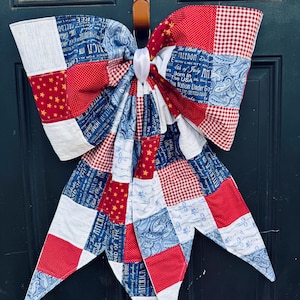 Puede incluir: Decoración de puerta en forma de lazo patriótico. Presenta un diseño de patchwork con telas rojas, blancas y azules. Algunas secciones tienen patrones de estrellas, estampados de cachemira y texto. El lazo está sujeto a una correa de cuero marrón.