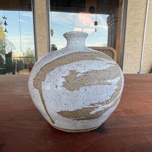 May include: A round ceramic vase with a narrow neck and a wide, bulbous body. The surface features a speckled, light tan clay base partially covered with thick, white, brushstroke-style glaze patterns that wrap around the exterior of the vessel.