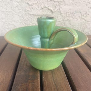 May include: A green ceramic candle holder with a curved handle. The candle holder has a bowl-like base with a raised center for the candle. The rim of the bowl has a brown accent. The candle holder is on a wooden surface.