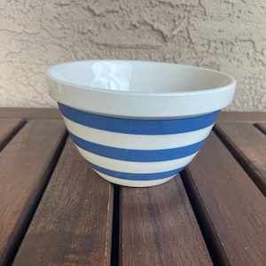 May include: A ceramic mixing bowl with a white interior and a blue and white striped exterior. The bowl is sitting on a wooden surface. The bowl is a kitchenware item.