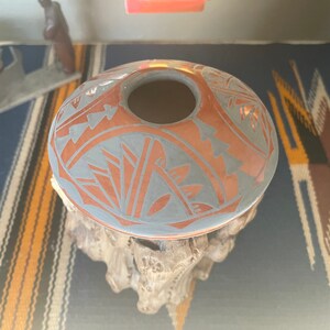 Puede incluir: Un jarrón de cerámica nativo americano gris y naranja con un diseño geométrico. El jarrón está sentado sobre un soporte de madera.