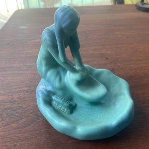 May include: A matte light blue figurine of a person kneeling and working at a pottery wheel on a circular base. The figure has long hair and is positioned over a small, flat, oval-shaped surface, with two small cylindrical objects resting nearby.