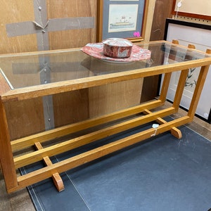May include: A wooden table with a glass top and two shelves. The table is made of light brown wood and has a simple design. A pink and white decorative object is on the top shelf.