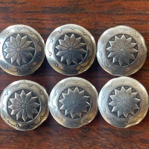 May include: A set of six circular silver-toned metal buttons, each featuring a central starburst or floral pattern with twelve petals. The outer edge of each button is decorated with a series of small, evenly spaced circular indentations.