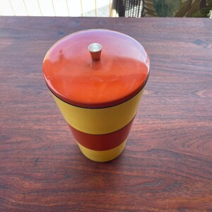 May include: A vintage, cylindrical container with a lid. The container is painted with horizontal bands of yellow, red, and orange. The lid is orange with a small silver knob. The container sits on a dark wooden surface.