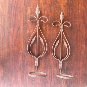 May include: Two ornate, vintage metal candle holders. Each features a teardrop-shaped design with a central leaf motif and a circular base. The candle holders are a rusty brown color and are set against a dark wood background.