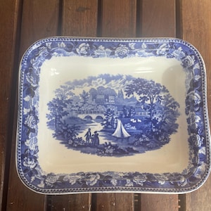 May include: A rectangular ceramic serving dish featuring a blue and white transferware pattern. The border displays a floral motif, while the center depicts a landscape scene with a bridge, a sailboat, trees, and figures near the water's edge.