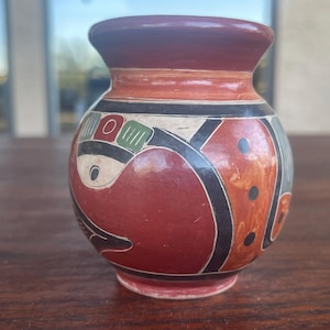 May include: A small, hand-painted ceramic vase with a rounded body and a narrow neck. The vase features a red base color with black, white, and orange geometric designs. The rim is a dark brown color.