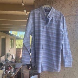 May include: A long-sleeved button-down shirt with a blue and white striped pattern. The shirt is hanging on a white hanger. The shirt has a button-down collar and buttoned cuffs. The shirt is made of a lightweight fabric.